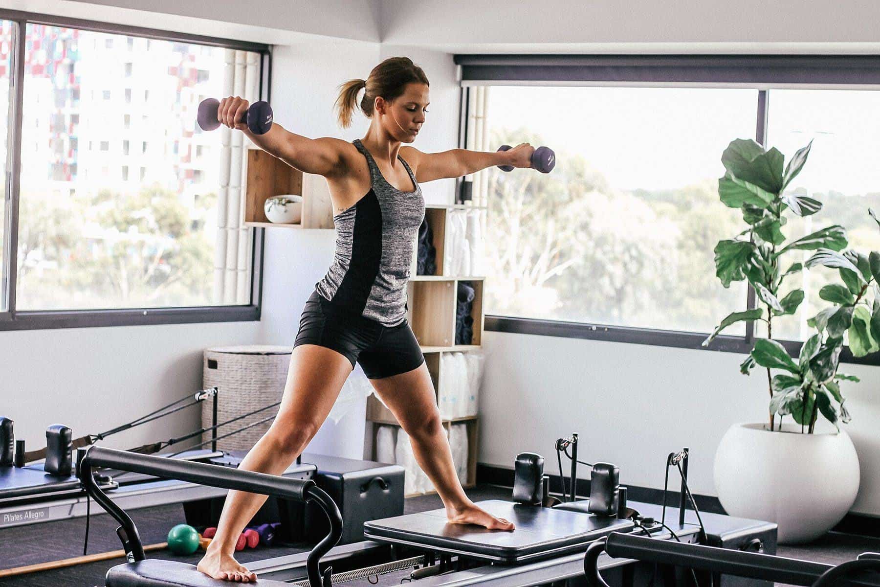 A woman standing on a reformer holding light hand weights in a bright Pilates studio