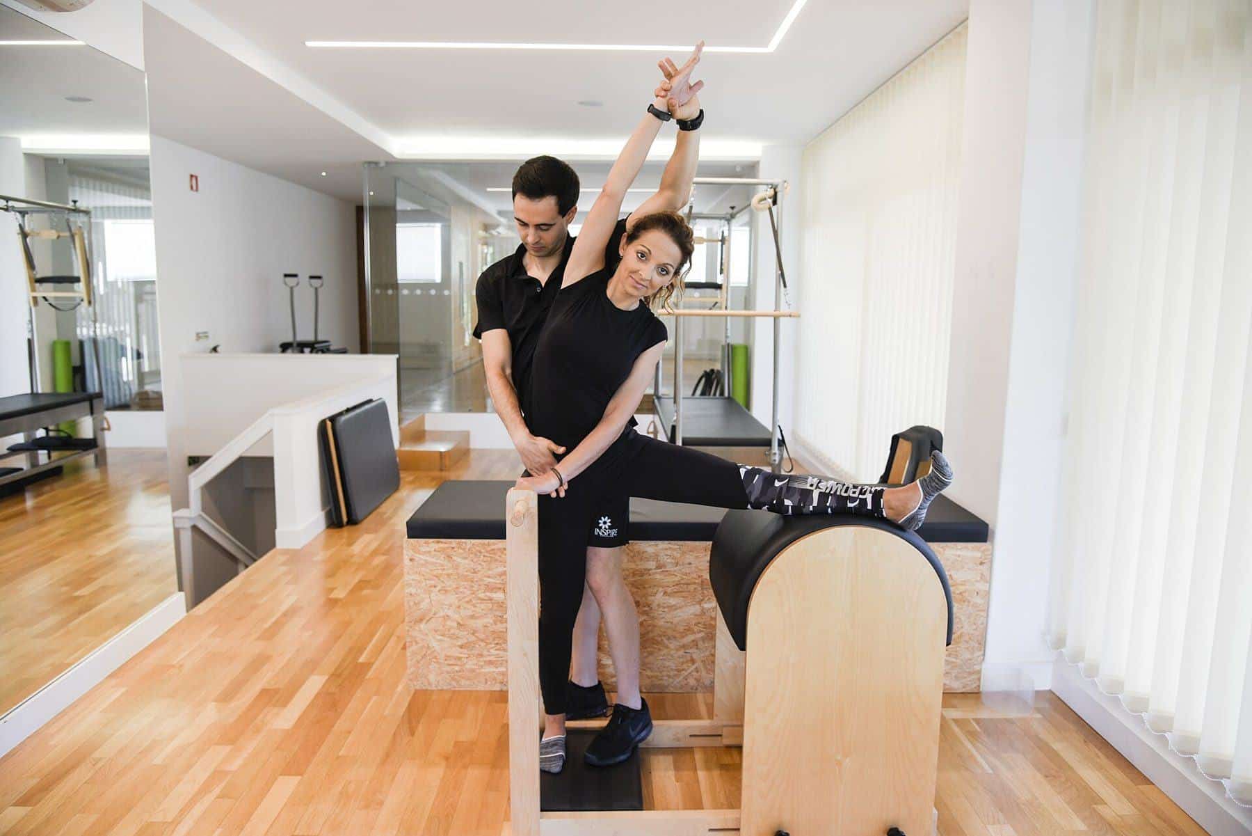 An instructor supporting a woman doing a stretch on a ladder barrel in a Pilates studio