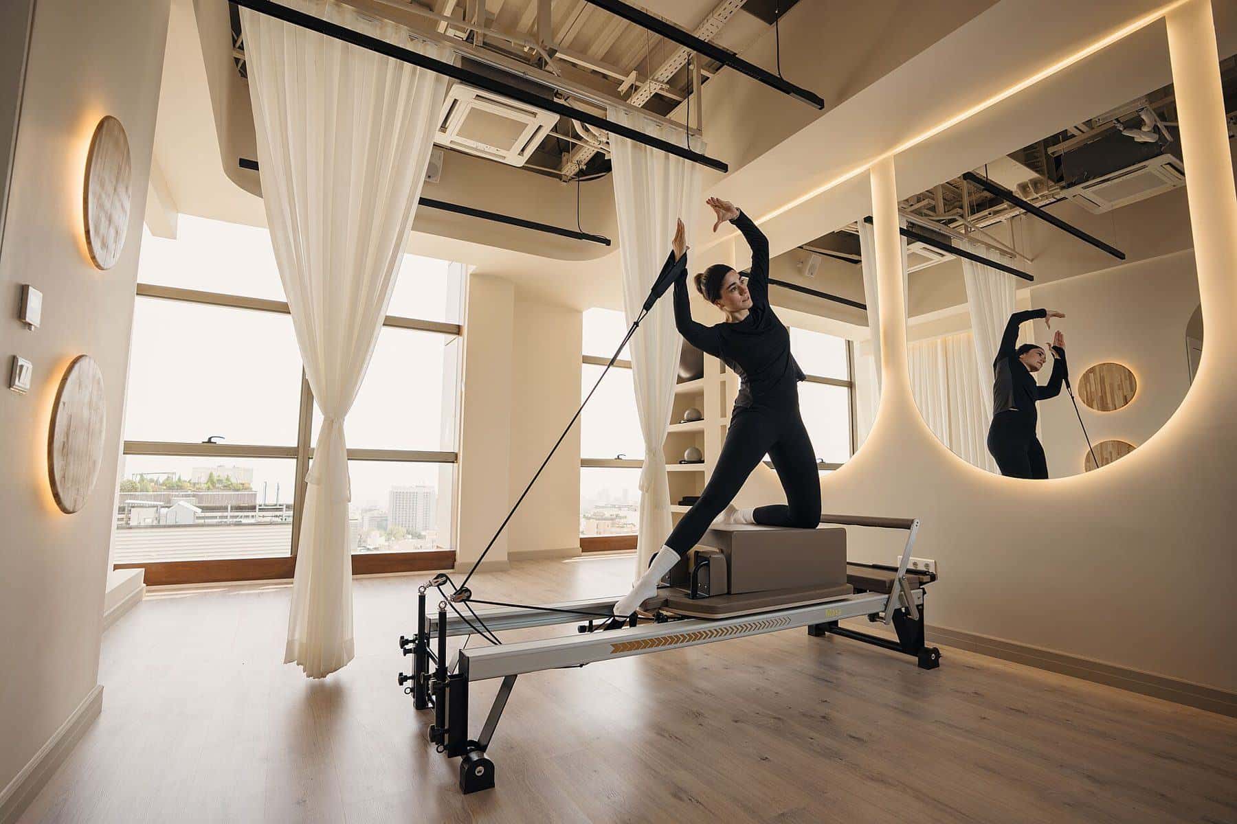 A woman standing on a reformer using the straps in a mirrored Pilates studio
