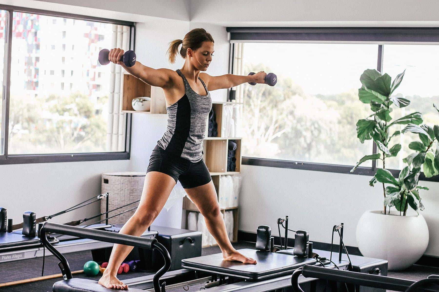 A woman standing on a reformer holding light weights in a bright studio with plants